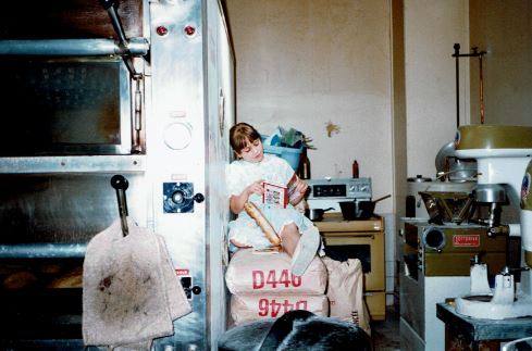 Irene sitting on flour bags alongside the original Italian Bakery equipment and snacking on a baguette circa 1988